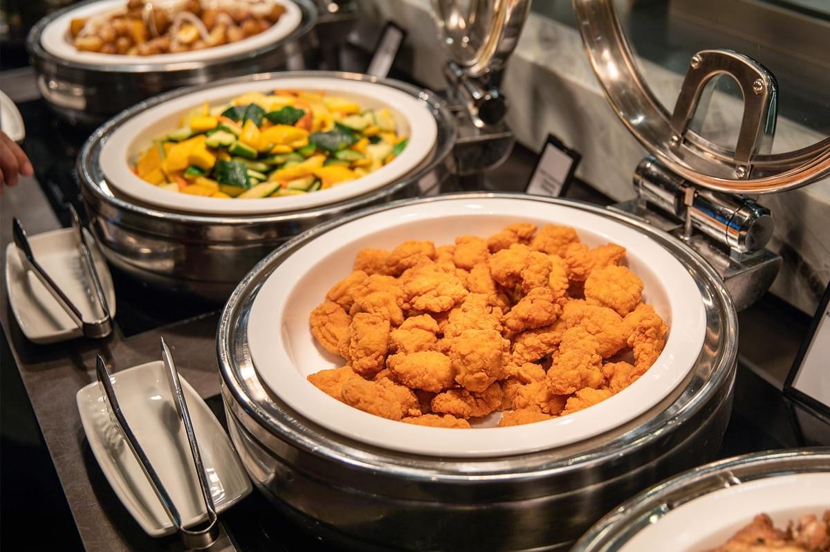 Buffet chafing dish filled with crispy fried chicken nuggets alongside a dish of roasted vegetables. Buffet chafing dish filled with crispy fried chicken nuggets alongside a dish of roasted vegetables.