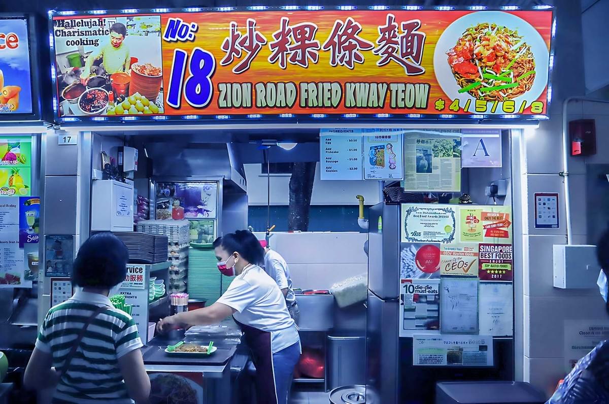 Hawker stall selling fried kway teow with signage reading "Zion Road Fried Kway Teow." The cook is busy preparing food, and customers are waiting. Hawker stall selling fried kway teow with signage reading "Zion Road Fried Kway Teow." The cook is busy preparing food, and customers are waiting.