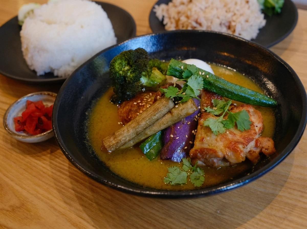 Bowl of Japanese soup curry topped with grilled chicken, okra, broccoli, and burdock root, served alongside rice and pickles. Bowl of Japanese soup curry topped with grilled chicken, okra, broccoli, and burdock root, served alongside rice and pickles.
