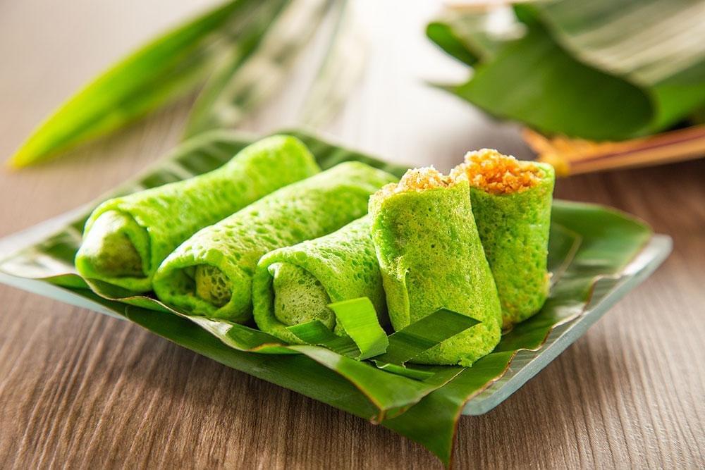 A plate of vibrant green, rolled pandan crepes, known as Kuih Ketayap, is beautifully arranged on top of a banana leaf. One of the crepes is cut in half, showcasing a rich filling of sweet, grated palm sugar coconut. A plate of vibrant green, rolled pandan crepes, known as Kuih Ketayap, is beautifully arranged on top of a banana leaf. One of the crepes is cut in half, showcasing a rich filling of sweet, grated palm sugar coconut.