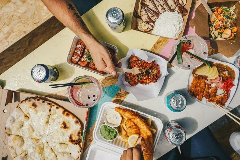 An overhead shot shows a vibrant, multi-colored outdoor table filled with an assortment of takeout food and drinks, including pizza, fish and chips, katsu curry, chicken wings, and a dessert crepe. Hands are visible reaching into the frame to enjoy the meal alongside canned beverages and cocktails garnished with fresh fruit. An overhead shot shows a vibrant, multi-colored outdoor table filled with an assortment of takeout food and drinks, including pizza, fish and chips, katsu curry, chicken wings, and a dessert crepe. Hands are visible reaching into the frame to enjoy the meal alongside canned beverages and cocktails garnished with fresh fruit.
