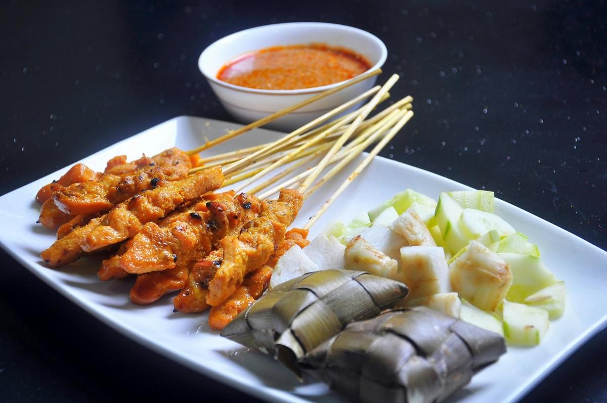 The image features a white rectangular plate filled with grilled chicken satay skewers, cubed rice cakes (ketupat), and chopped cucumber slices against a dark background. In the background, a small white bowl is filled with a thick, reddish-brown peanut dipping sauce. The image features a white rectangular plate filled with grilled chicken satay skewers, cubed rice cakes (ketupat), and chopped cucumber slices against a dark background. In the background, a small white bowl is filled with a thick, reddish-brown peanut dipping sauce.