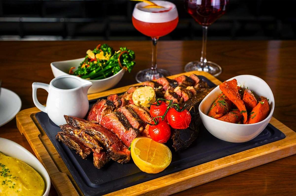 A wooden board holds sliced grilled steak topped with butter and roasted tomatoes, beside a bowl of glazed carrots. Accompanied by sides and vibrant cocktails. A wooden board holds sliced grilled steak topped with butter and roasted tomatoes, beside a bowl of glazed carrots. Accompanied by sides and vibrant cocktails.