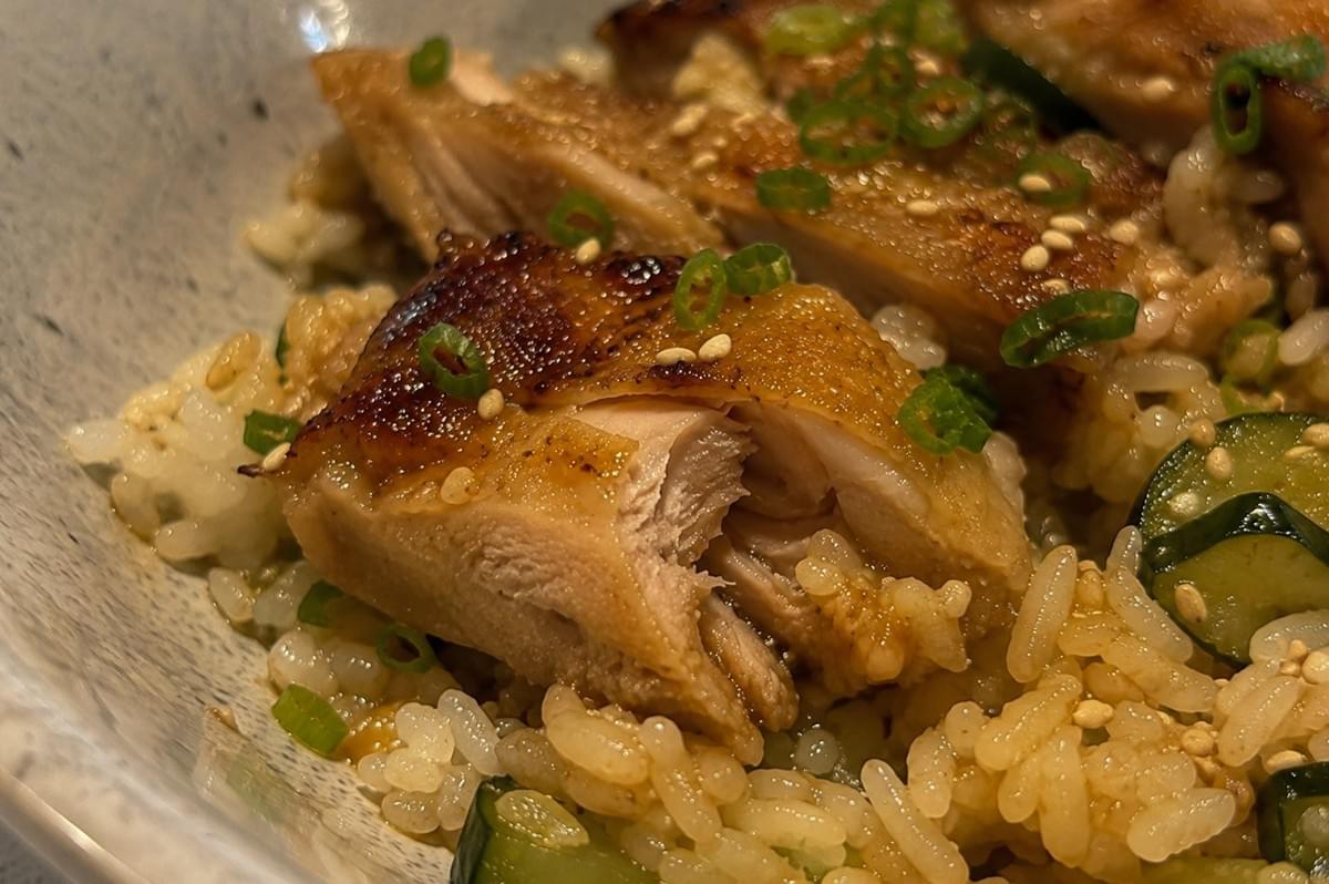 A close-up view of a bowl containing perfectly cooked, sliced chicken thighs with crispy, browned skin. The meat is garnished with a generous sprinkling of finely chopped green onions and toasted sesame seeds, resting atop a bed of rice mixed with thinly sliced, marinated cucumbers. A close-up view of a bowl containing perfectly cooked, sliced chicken thighs with crispy, browned skin. The meat is garnished with a generous sprinkling of finely chopped green onions and toasted sesame seeds, resting atop a bed of rice mixed with thinly sliced, marinated cucumbers.
