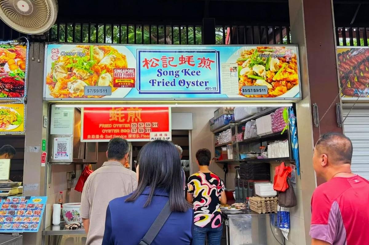 A small food stall named "Song Kee Fried Oyster" with a colorful signboard showing oyster dishes. Three people wait in line, conveying a busy, vibrant atmosphere. A small food stall named "Song Kee Fried Oyster" with a colorful signboard showing oyster dishes. Three people wait in line, conveying a busy, vibrant atmosphere.