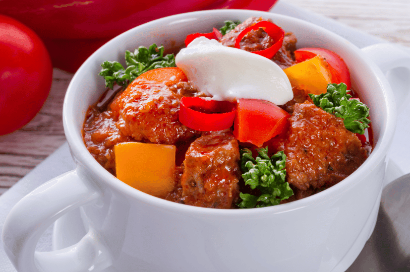 A comforting white bowl of chunky Hungarian goulash topped with a dollop of white sour cream, fresh bell pepper slices, and parsley. A comforting white bowl of chunky Hungarian goulash topped with a dollop of white sour cream, fresh bell pepper slices, and parsley.