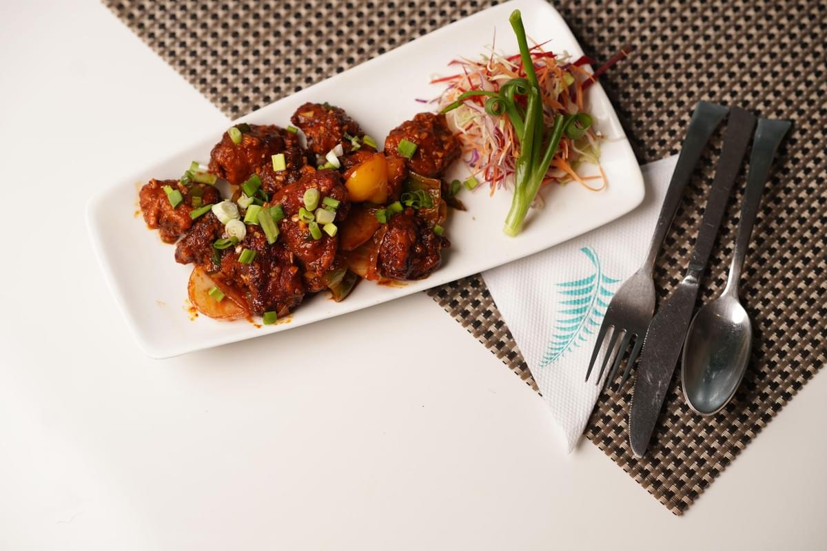 The image features a rectangular white plate holding a serving of saucy, dark red-brown Manchurian or stir-fried chicken pieces garnished with chopped green onions. Beside the main dish is a colorful side salad with shredded cabbage, carrots, and whole green onion stalks, arranged on a patterned placemat next to a fork, knife, and spoon. The image features a rectangular white plate holding a serving of saucy, dark red-brown Manchurian or stir-fried chicken pieces garnished with chopped green onions. Beside the main dish is a colorful side salad with shredded cabbage, carrots, and whole green onion stalks, arranged on a patterned placemat next to a fork, knife, and spoon.