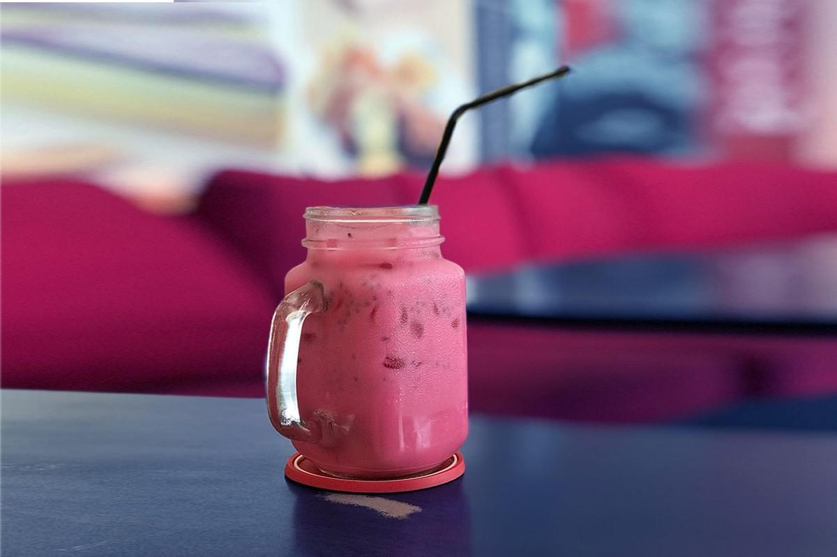 A pink iced drink in a mason jar sits on a glossy blue table. It has a black straw and red coaster. The background is a blurred, vibrant pink sofa. A pink iced drink in a mason jar sits on a glossy blue table. It has a black straw and red coaster. The background is a blurred, vibrant pink sofa.