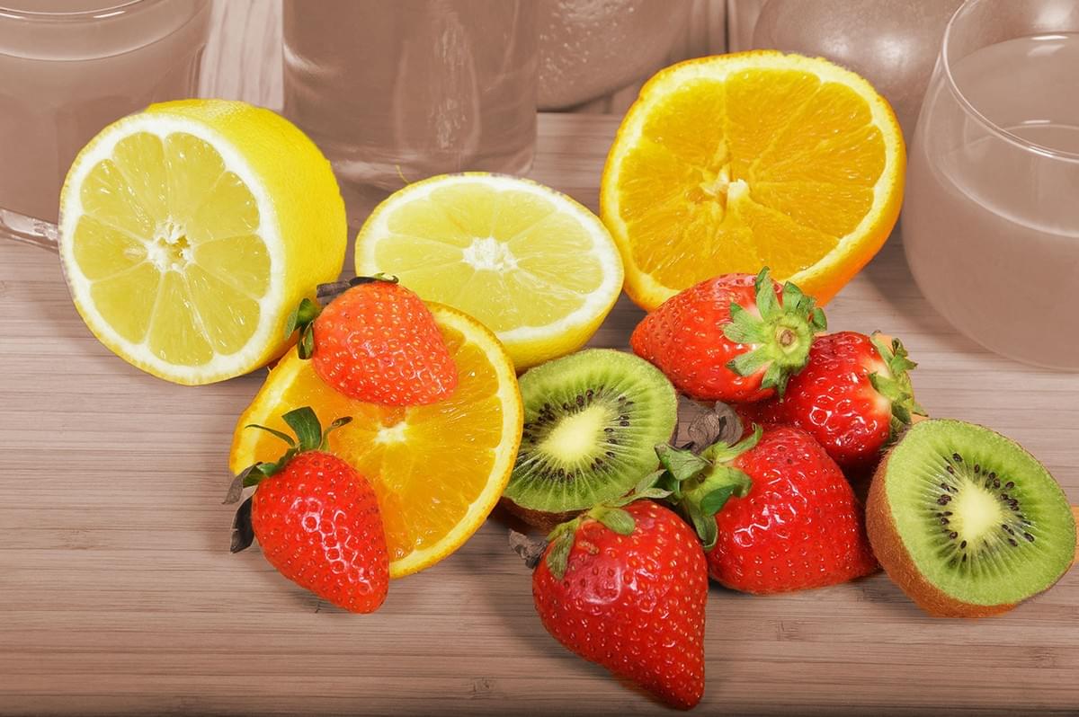 A wooden table displays vibrant strawberries, halved lemons and oranges, and sliced kiwis. In the background are glasses of a pale beverage, creating a fresh, summery feel. A wooden table displays vibrant strawberries, halved lemons and oranges, and sliced kiwis. In the background are glasses of a pale beverage, creating a fresh, summery feel.