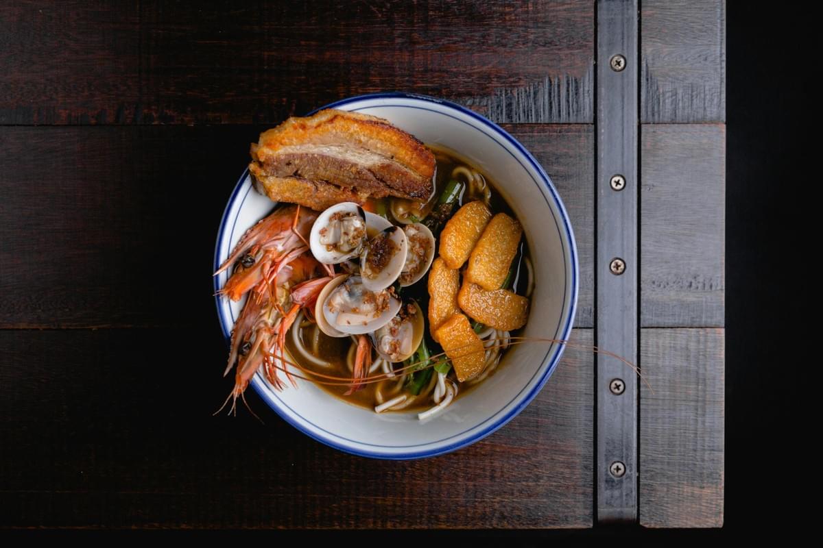 A top-down view shows a bowl of noodle soup generously topped with crispy pork belly, whole head-on shrimp, clams, and fried tofu puffs. The blue-rimmed white bowl rests on a dark, rustic wooden tabletop. A top-down view shows a bowl of noodle soup generously topped with crispy pork belly, whole head-on shrimp, clams, and fried tofu puffs. The blue-rimmed white bowl rests on a dark, rustic wooden tabletop.