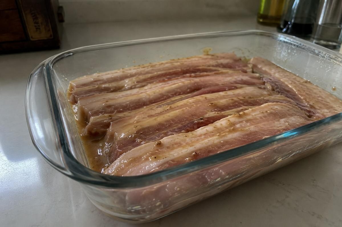 A close-up, angled shot shows several thick, raw strips of pork belly marinating in a shallow, clear glass baking dish on a light-colored kitchen countertop. The pork belly slices are laid out flat in a neat row, coated in a light-brown, speckled liquid marinade that pool slightly at the bottom of the dish. The background is softly out of focus, showing a glimpse of a wooden block on the left and blurred kitchen bottles on the right under soft, natural lighting. A close-up, angled shot shows several thick, raw strips of pork belly marinating in a shallow, clear glass baking dish on a light-colored kitchen countertop. The pork belly slices are laid out flat in a neat row, coated in a light-brown, speckled liquid marinade that pool slightly at the bottom of the dish. The background is softly out of focus, showing a glimpse of a wooden block on the left and blurred kitchen bottles on the right under soft, natural lighting.