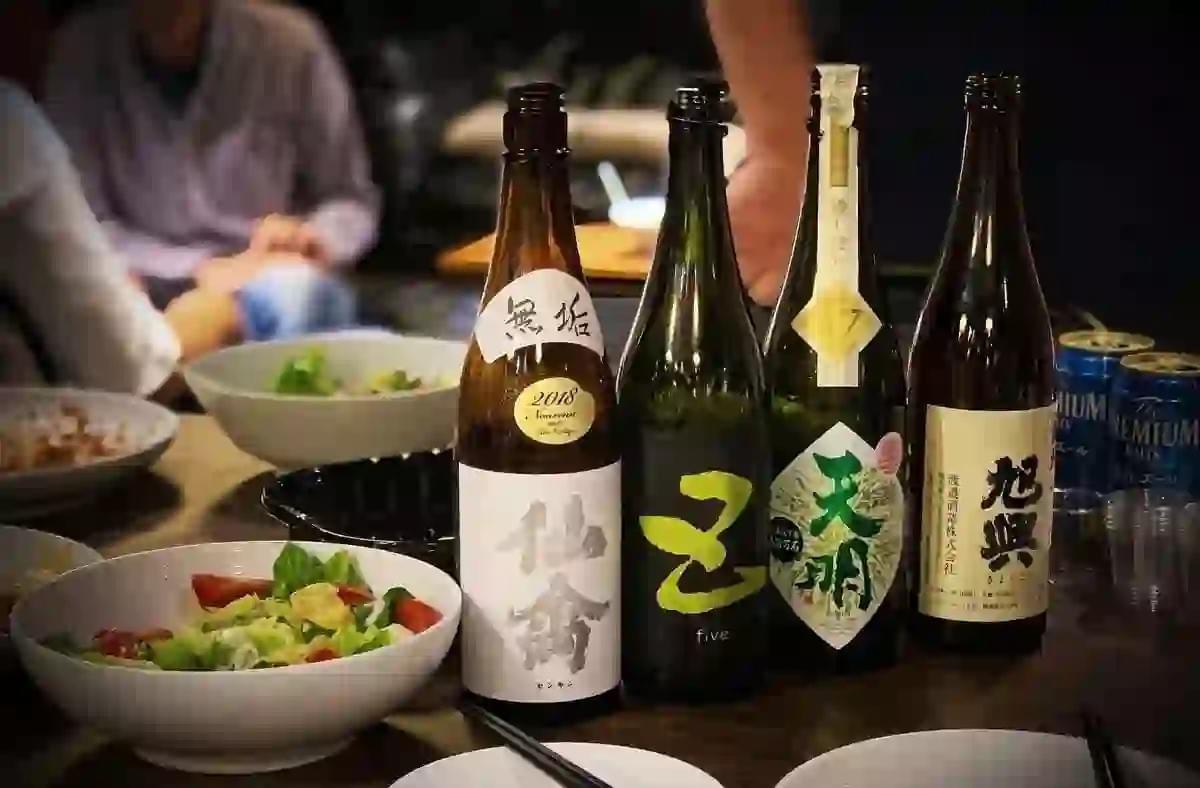 Four bottles of Japanese sake with distinct labels stand on a table alongside fresh salad bowls and blue cans of Suntory beer. The blurred background shows a cozy izakaya setting with people enjoying a meal in soft, warm lighting. Four bottles of Japanese sake with distinct labels stand on a table alongside fresh salad bowls and blue cans of Suntory beer. The blurred background shows a cozy izakaya setting with people enjoying a meal in soft, warm lighting.
