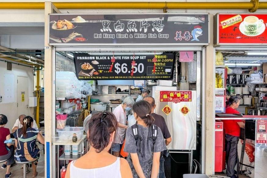 A storefront for "Mei Xiang Black & White Fish Soup" displays a prominent menu board advertising a set meal for $6.50. Customers are seen waiting in line in front of the food stall. A storefront for "Mei Xiang Black & White Fish Soup" displays a prominent menu board advertising a set meal for $6.50. Customers are seen waiting in line in front of the food stall.