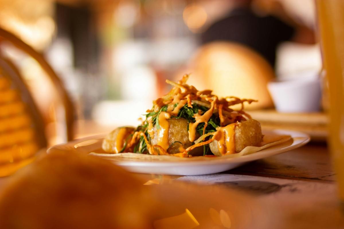 A close-up view of a taco filled with crispy fried bites, fresh green herbs, and crisp toppings, drizzled with a creamy orange sauce. The plate sits on a wooden restaurant table under warm, golden lighting with a softly blurred background. A close-up view of a taco filled with crispy fried bites, fresh green herbs, and crisp toppings, drizzled with a creamy orange sauce. The plate sits on a wooden restaurant table under warm, golden lighting with a softly blurred background.