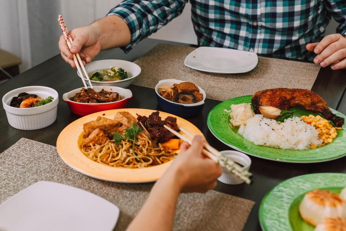 Two people sit at a table filled with various Asian dishes, including noodles, rice, chicken, and side dishes in small bowls. They use chopsticks to reach for and share the food laid out across colorful plates and placemats. Two people sit at a table filled with various Asian dishes, including noodles, rice, chicken, and side dishes in small bowls. They use chopsticks to reach for and share the food laid out across colorful plates and placemats.