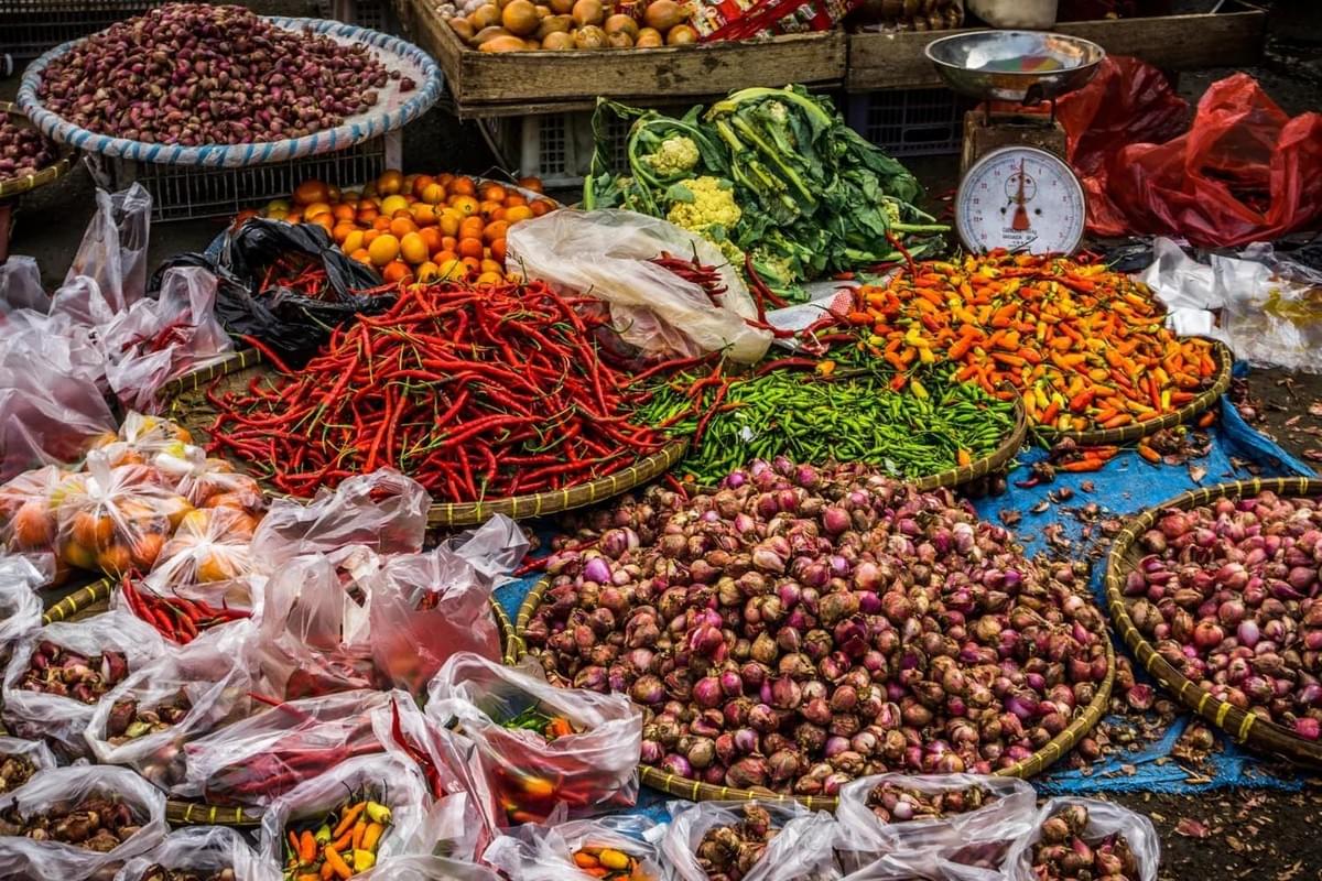 A vibrant outdoor market stall displays an abundance of fresh produce, including large mounds of red and green chilies alongside piles of shallots and tomatoes. Woven baskets and plastic bags overflow with colorful vegetables, while a mechanical weighing scale sits ready for customers in the background. A vibrant outdoor market stall displays an abundance of fresh produce, including large mounds of red and green chilies alongside piles of shallots and tomatoes. Woven baskets and plastic bags overflow with colorful vegetables, while a mechanical weighing scale sits ready for customers in the background.