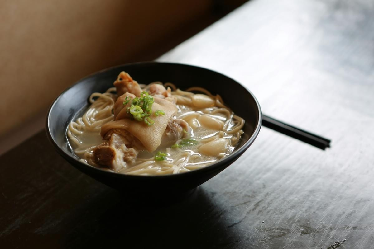 A steaming black bowl of noodle soup featuring a large piece of pork trotter, garlic cloves, and a garnish of chopped green onions sits on a dark wooden table. A pair of black chopsticks rests beside the bowl on the glossy, reflective tabletop. A steaming black bowl of noodle soup featuring a large piece of pork trotter, garlic cloves, and a garnish of chopped green onions sits on a dark wooden table. A pair of black chopsticks rests beside the bowl on the glossy, reflective tabletop.