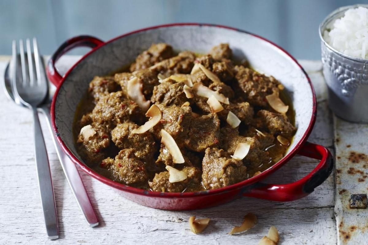 This dish features tender chunks of slow-cooked meat coated in a thick, aromatic curry sauce and topped with toasted coconut flakes. Served in a rustic red-handled pan alongside a bowl of white rice, the meal sits on a weathered wooden surface for a cozy, homemade feel. This dish features tender chunks of slow-cooked meat coated in a thick, aromatic curry sauce and topped with toasted coconut flakes. Served in a rustic red-handled pan alongside a bowl of white rice, the meal sits on a weathered wooden surface for a cozy, homemade feel.