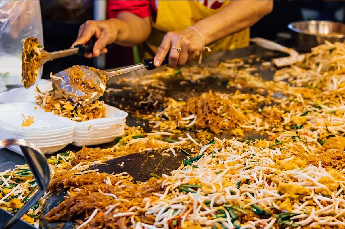 Medium close-up action shot of a Thai street food vendor cooking and serving traditional pad thai on a hot flat-top wok, with stir-fried rice noodles, bean sprouts, and eggs in a busy Bangkok street kitchen. Medium close-up action shot of a Thai street food vendor cooking and serving traditional pad thai on a hot flat-top wok, with stir-fried rice noodles, bean sprouts, and eggs in a busy Bangkok street kitchen.