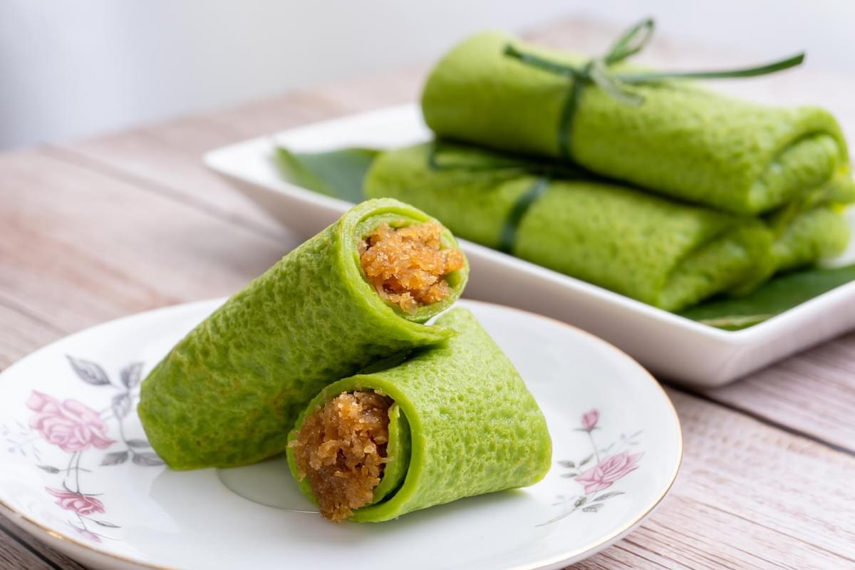 A plate in the foreground holds a bright green, pandan-infused crepe rolled around a sweet, grated coconut filling and cut in half to show the inside. In the background, more intact crepes are neatly stacked on a rectangular dish, with some tied with pandan leaves for decoration. A plate in the foreground holds a bright green, pandan-infused crepe rolled around a sweet, grated coconut filling and cut in half to show the inside. In the background, more intact crepes are neatly stacked on a rectangular dish, with some tied with pandan leaves for decoration.