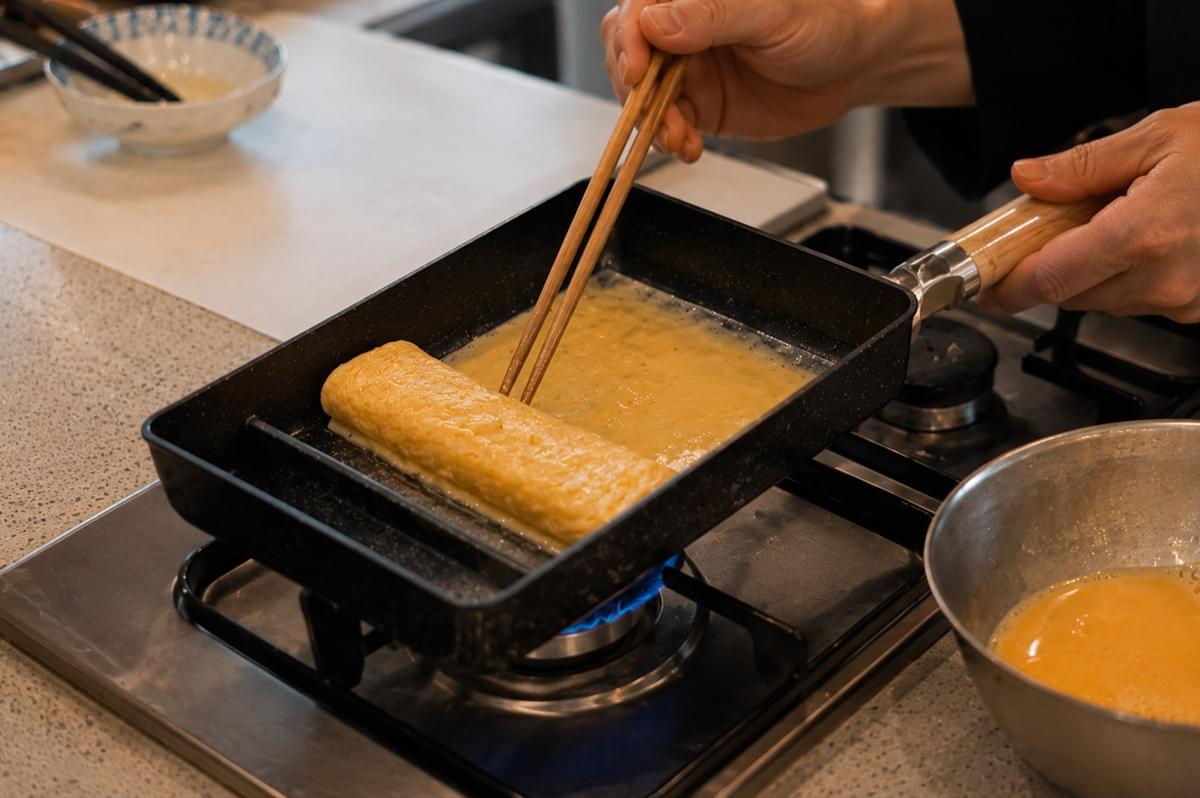 A close-up view shows wooden chopsticks guiding the edge of a delicate egg layer as it cooks in a non-stick rectangular pan, gradually building the rolled tamagoyaki over a blue gas flame. A close-up view shows wooden chopsticks guiding the edge of a delicate egg layer as it cooks in a non-stick rectangular pan, gradually building the rolled tamagoyaki over a blue gas flame.