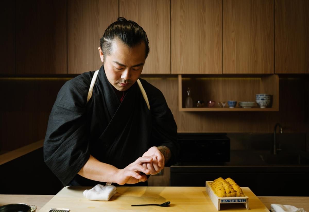 A sushi chef in a black kimono meticulously shapes a piece of nigiri at a clean, light-wood counter. Beside him, a wooden tray of fresh, golden uni sits ready for preparation against a backdrop of modern minimalist cabinetry. A sushi chef in a black kimono meticulously shapes a piece of nigiri at a clean, light-wood counter. Beside him, a wooden tray of fresh, golden uni sits ready for preparation against a backdrop of modern minimalist cabinetry.