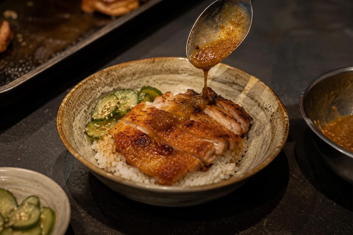 A silver spoon held over a rustic, speckled ceramic bowl pours a glossy, brown sauce onto slices of golden-brown, pan-seared chicken. The chicken rests on a bed of fluffy white rice, accompanied by a small side of thinly sliced cucumbers sprinkled with sesame seeds, all set against a dark, moody background. A silver spoon held over a rustic, speckled ceramic bowl pours a glossy, brown sauce onto slices of golden-brown, pan-seared chicken. The chicken rests on a bed of fluffy white rice, accompanied by a small side of thinly sliced cucumbers sprinkled with sesame seeds, all set against a dark, moody background.