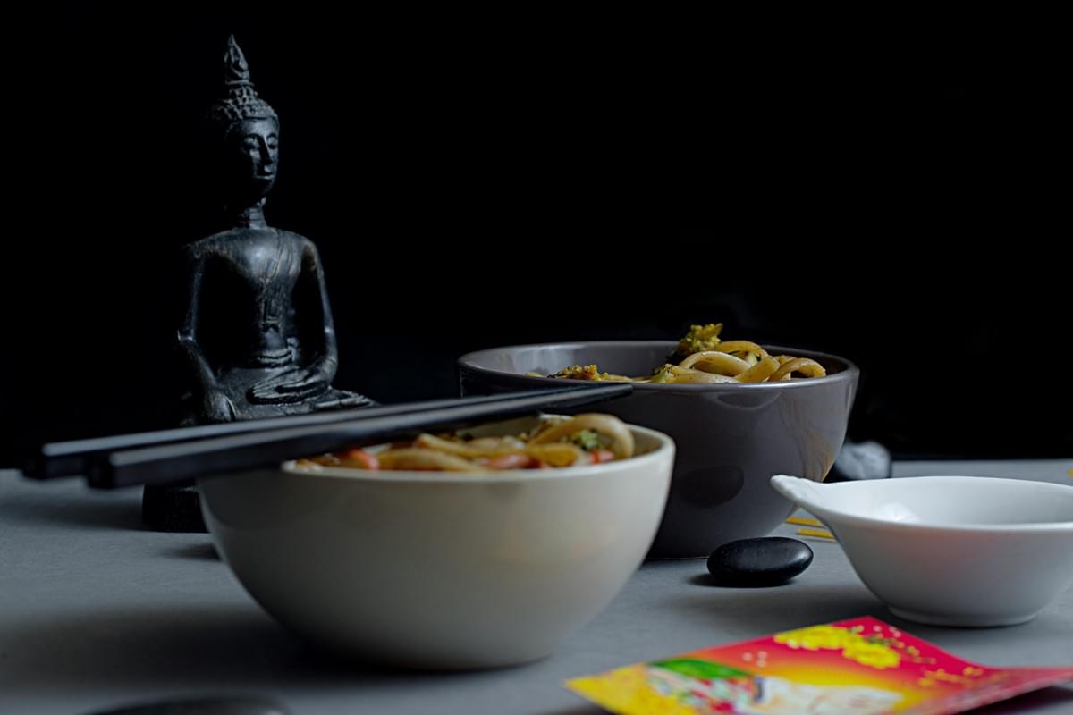 A small dark Buddha statue sits in the background against a black backdrop next to two bowls of stir-fried noodles with chopsticks. In the foreground, a white dipping bowl, polished black stones, and a colorful card rest on a gray surface. A small dark Buddha statue sits in the background against a black backdrop next to two bowls of stir-fried noodles with chopsticks. In the foreground, a white dipping bowl, polished black stones, and a colorful card rest on a gray surface.