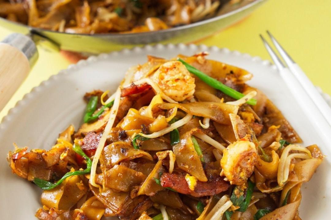 This image displays a plate of Penang Char Kuey Teow, showing wide rice noodles stir-fried with shrimp, bean sprouts, and Chinese sausage. The dish is presented on a white plate with a wok visible in the background against a yellow surface. This image displays a plate of Penang Char Kuey Teow, showing wide rice noodles stir-fried with shrimp, bean sprouts, and Chinese sausage. The dish is presented on a white plate with a wok visible in the background against a yellow surface.