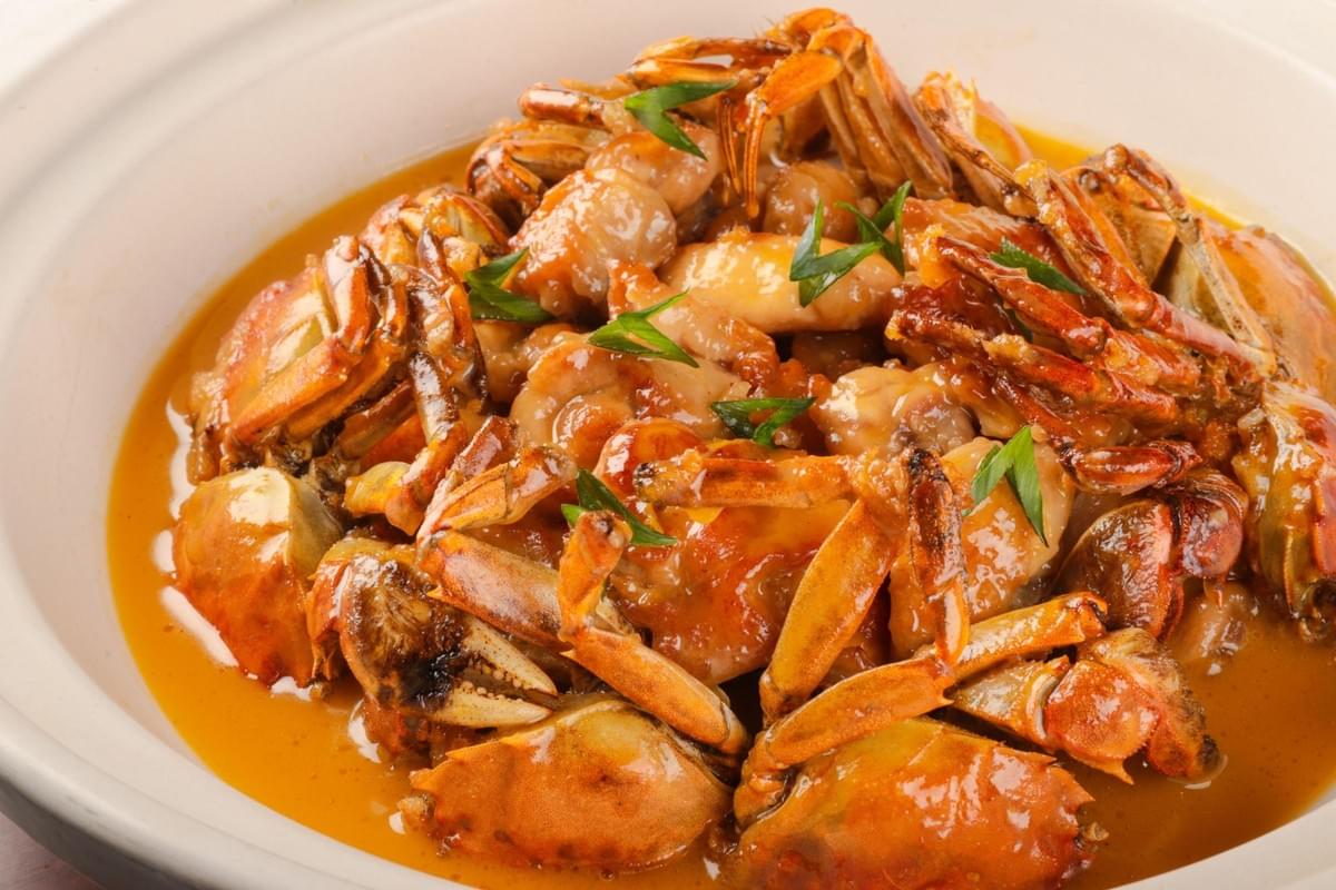 A high-angle view shows a ceramic bowl filled with cooked small crabs and chicken pieces coated in a rich, savory orange sauce. The dish is garnished with fresh sliced green scallions scattered across the top. A high-angle view shows a ceramic bowl filled with cooked small crabs and chicken pieces coated in a rich, savory orange sauce. The dish is garnished with fresh sliced green scallions scattered across the top.