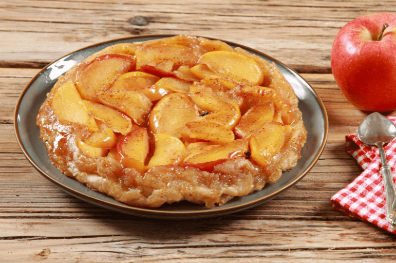 A golden, glossy Tarte Tatin on a blue plate, set on a wooden table next to a fresh red apple and a spoon on a red-and-white checkered napkin. A golden, glossy Tarte Tatin on a blue plate, set on a wooden table next to a fresh red apple and a spoon on a red-and-white checkered napkin.