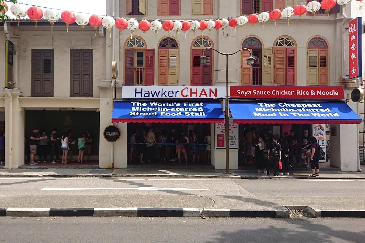The exterior of Hawker CHAN, a restaurant selling soya sauce chicken rice and noodles, features blue awnings declaring it as the world's first Michelin-starred street food stall. Red and white lanterns hang across the front of the traditional heritage building while a line of customers waits outside. The exterior of Hawker CHAN, a restaurant selling soya sauce chicken rice and noodles, features blue awnings declaring it as the world's first Michelin-starred street food stall. Red and white lanterns hang across the front of the traditional heritage building while a line of customers waits outside.