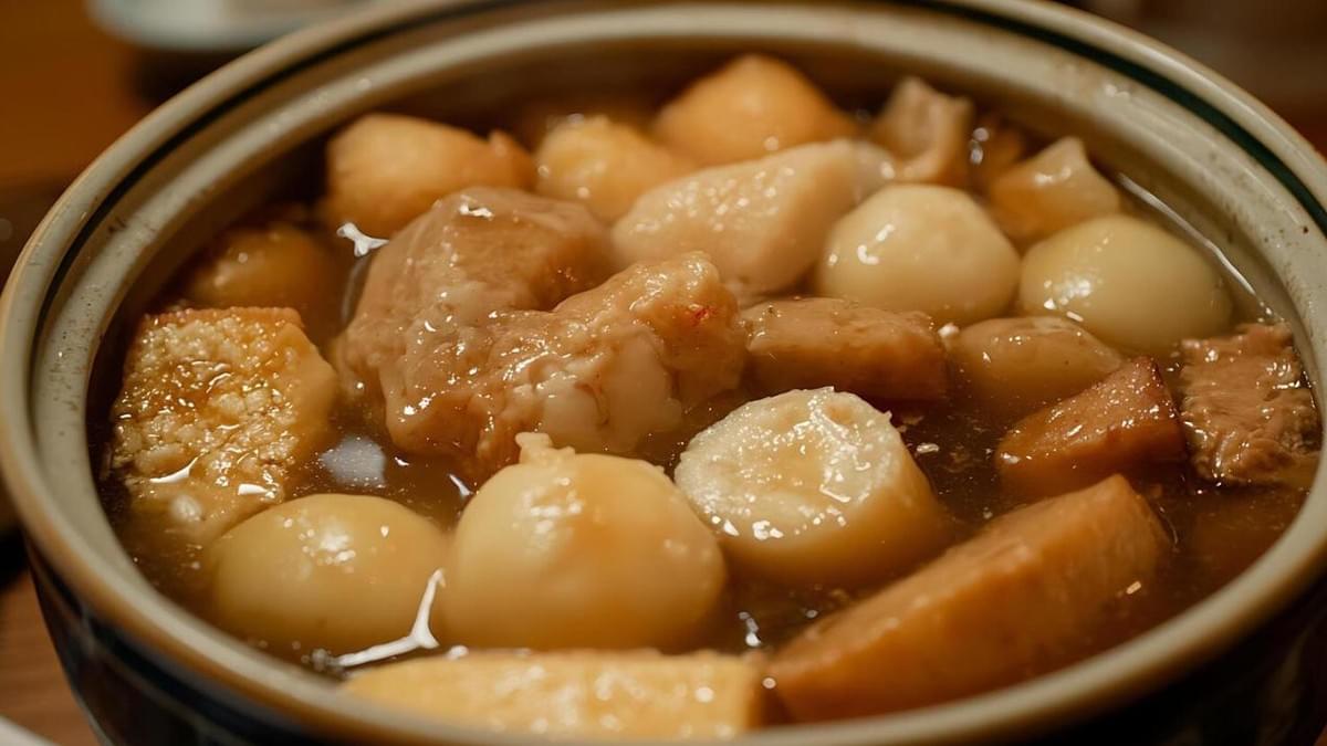 A ceramic bowl is filled with a glossy, amber-colored broth containing round white dumplings, chunks of root vegetables, and pieces of textured ingredients that resemble fried dough or tofu. The warm lighting highlights the glistening surfaces of the hearty contents, which are packed tightly within the rimmed container. A ceramic bowl is filled with a glossy, amber-colored broth containing round white dumplings, chunks of root vegetables, and pieces of textured ingredients that resemble fried dough or tofu. The warm lighting highlights the glistening surfaces of the hearty contents, which are packed tightly within the rimmed container.