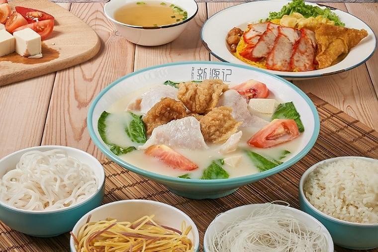 A large bowl of milky fish soup with fried fish pieces, leafy greens, tomatoes, and tofu sits at the center of a light wooden table. Surrounding it are side bowls of various noodles, rice, clear broth, char siu noodles, and sliced tofu with tomatoes. A large bowl of milky fish soup with fried fish pieces, leafy greens, tomatoes, and tofu sits at the center of a light wooden table. Surrounding it are side bowls of various noodles, rice, clear broth, char siu noodles, and sliced tofu with tomatoes.
