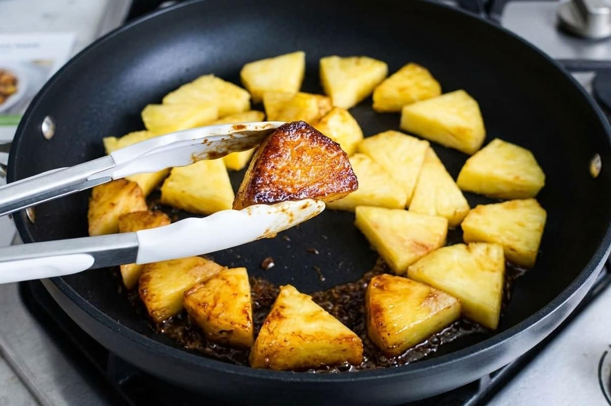 Tongs lifting a caramelized pineapple chunk from a black skillet filled with browning pineapple pieces Tongs lifting a caramelized pineapple chunk from a black skillet filled with browning pineapple pieces