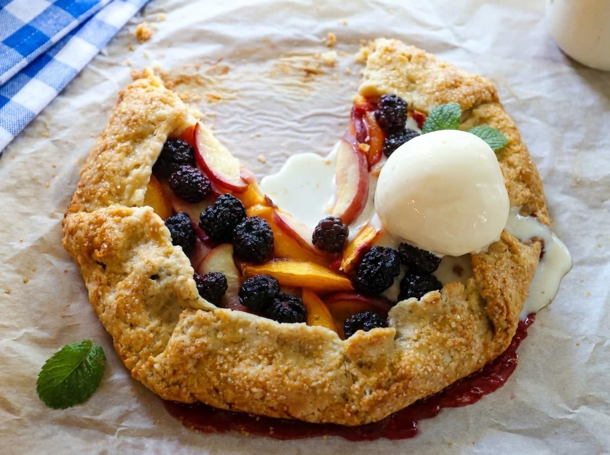 A rustic fruit galette filled with peaches and blackberries rests on parchment paper, topped with a scoop of melting vanilla ice cream and garnished with fresh mint leaves. The dessert is photographed in natural light, creating a fresh and inviting homemade bakery feel. A rustic fruit galette filled with peaches and blackberries rests on parchment paper, topped with a scoop of melting vanilla ice cream and garnished with fresh mint leaves. The dessert is photographed in natural light, creating a fresh and inviting homemade bakery feel.