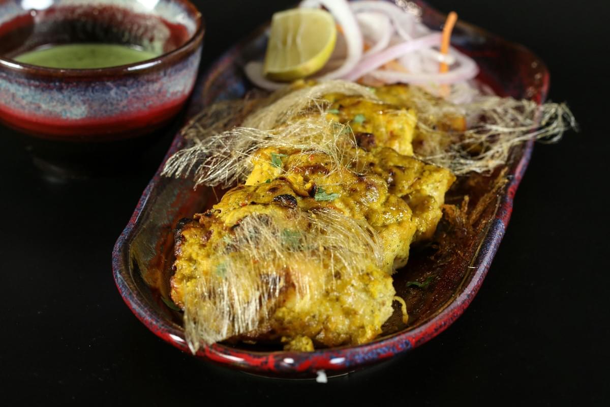 This appetizing high-angle shot presents a platter of golden-yellow grilled chicken pieces, uniquely garnished with delicate, hair-like threads that add an intriguing texture. The dish is accompanied by a side of fresh onion rings, a lime wedge, and a small bowl of green dipping sauce, all set against a sleek black background that highlights the vibrant food colors. This appetizing high-angle shot presents a platter of golden-yellow grilled chicken pieces, uniquely garnished with delicate, hair-like threads that add an intriguing texture. The dish is accompanied by a side of fresh onion rings, a lime wedge, and a small bowl of green dipping sauce, all set against a sleek black background that highlights the vibrant food colors.