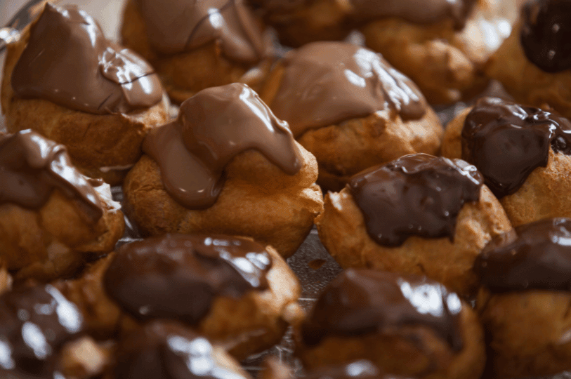 A close-up of a batch of classic profiteroles, featuring golden choux pastry puffs generously topped with a smooth, thick chocolate glaze. A close-up of a batch of classic profiteroles, featuring golden choux pastry puffs generously topped with a smooth, thick chocolate glaze.
