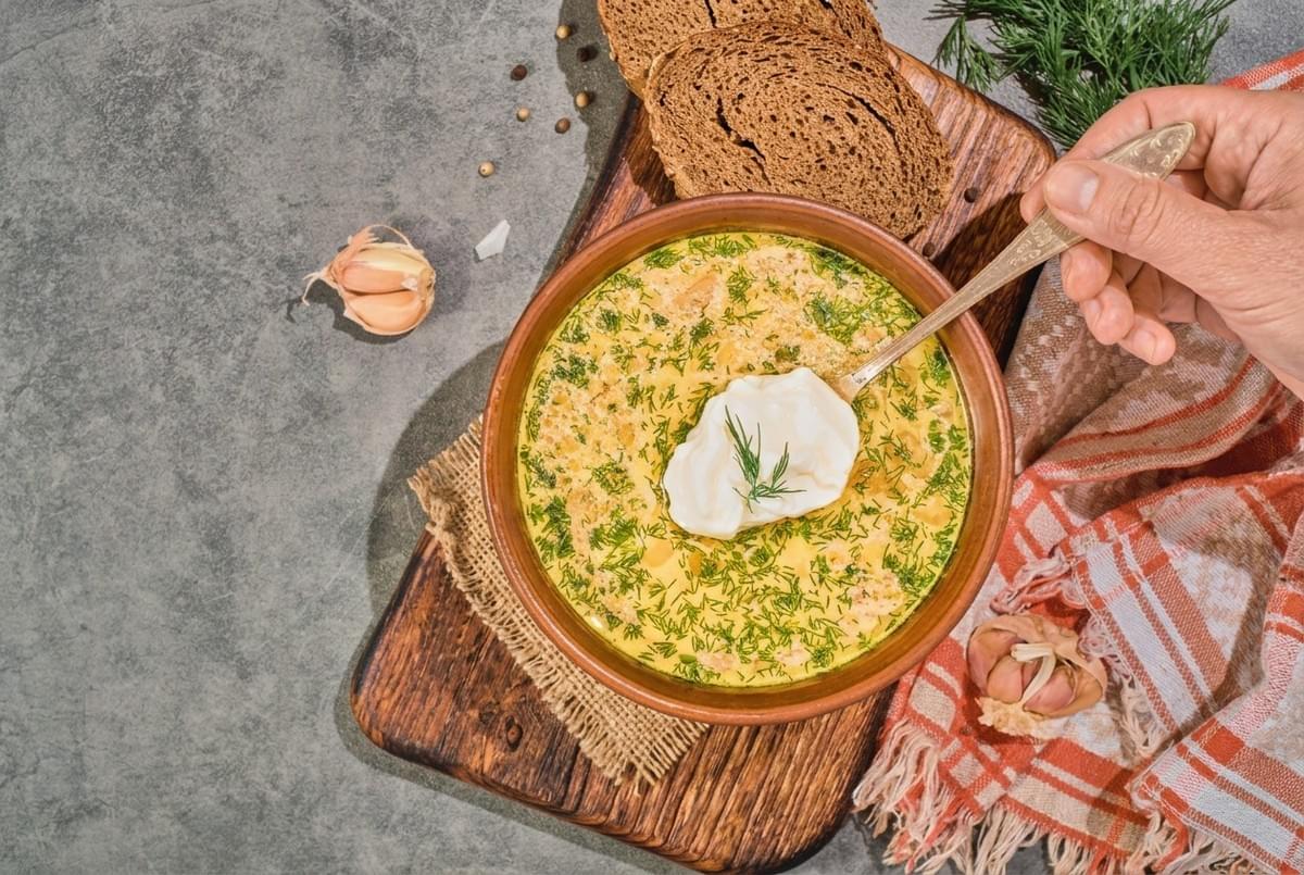 Clay bowl of yellow dill soup topped with sour cream, served alongside rye bread slices. Clay bowl of yellow dill soup topped with sour cream, served alongside rye bread slices.