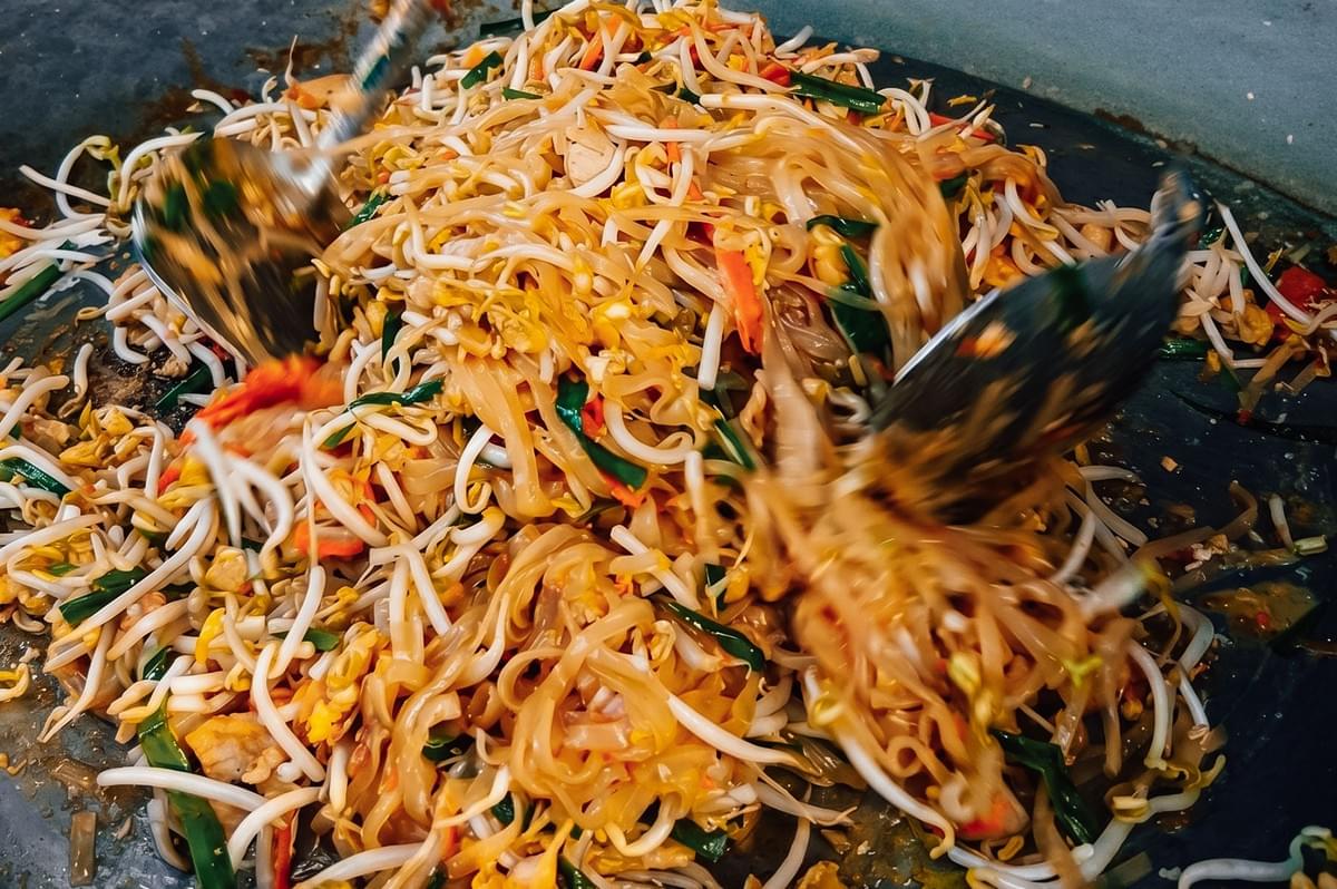 Dynamic overhead close-up shot of pad thai being tossed in a wok, showing authentic Thai stir-fried rice noodles with bean sprouts, eggs, and garlic chives creating wok hei flavor. Dynamic overhead close-up shot of pad thai being tossed in a wok, showing authentic Thai stir-fried rice noodles with bean sprouts, eggs, and garlic chives creating wok hei flavor.