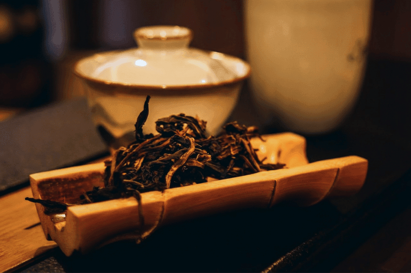 A macro shot focusing on dark, premium loose tea leaves resting in a curved bamboo chahe display vessel, with a soft-focus white ceramic gaiwan in the background at the Tea Room by Ki-setsu. A macro shot focusing on dark, premium loose tea leaves resting in a curved bamboo chahe display vessel, with a soft-focus white ceramic gaiwan in the background at the Tea Room by Ki-setsu.