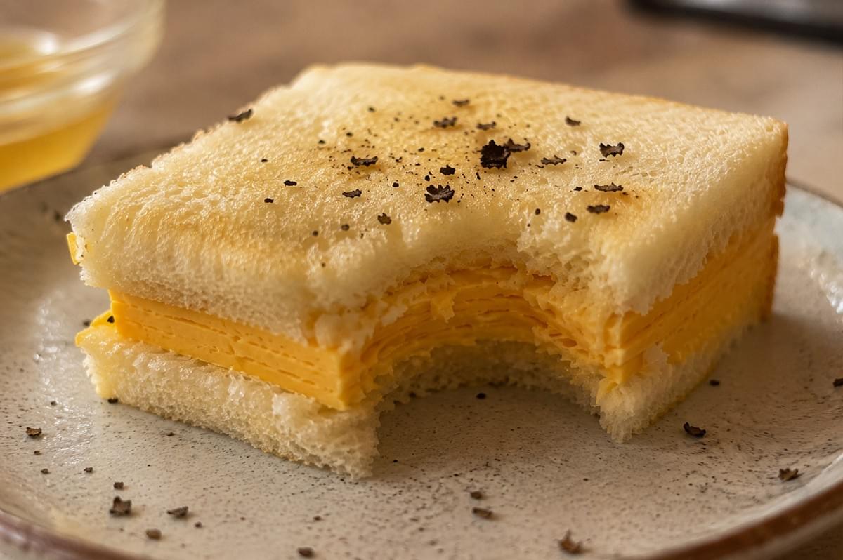 A close-up, tempting view of a single Tamago Sando half shows a large bite taken out, revealing the dense, delicate horizontal layers of the rolled dashi omelet inside. The bread is soft and airy, with its surface lightly glistening from a drizzle of truffle oil and a generous sprinkling of earthy black truffle bits. The cross-section highlights the moist, custard-like texture of the egg, while the soft lighting and shallow depth of field focus entirely on the rich textures of the sandwich, making the "elevated" nature of this classic comfort food visceral. A close-up, tempting view of a single Tamago Sando half shows a large bite taken out, revealing the dense, delicate horizontal layers of the rolled dashi omelet inside. The bread is soft and airy, with its surface lightly glistening from a drizzle of truffle oil and a generous sprinkling of earthy black truffle bits. The cross-section highlights the moist, custard-like texture of the egg, while the soft lighting and shallow depth of field focus entirely on the rich textures of the sandwich, making the "elevated" nature of this classic comfort food visceral.