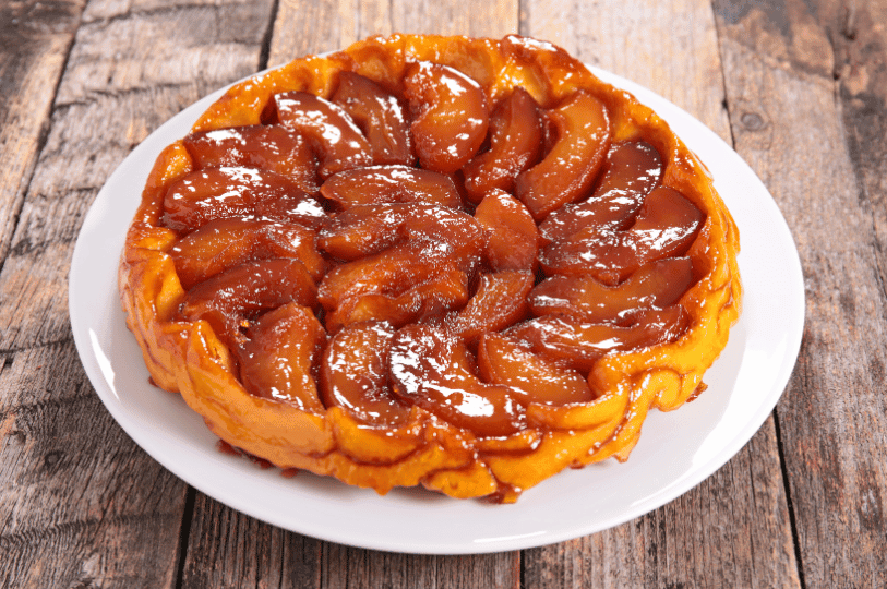 A freshly baked classic Tarte Tatin showcasing tightly packed, rich amber caramelized apples on a simple white plate over a rustic wooden background. A freshly baked classic Tarte Tatin showcasing tightly packed, rich amber caramelized apples on a simple white plate over a rustic wooden background.