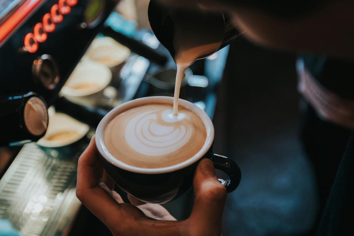 A barista pours steamed milk from a metal pitcher into a cup of espresso, creating decorative latte art on the surface. An espresso machine with glowing orange lights is visible in the background. A barista pours steamed milk from a metal pitcher into a cup of espresso, creating decorative latte art on the surface. An espresso machine with glowing orange lights is visible in the background.