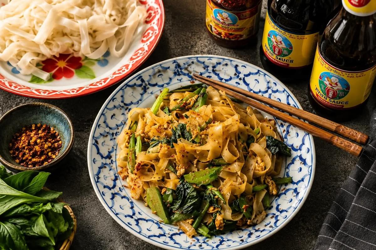 Top-down view of a blue patterned plate of stir-fried Pad See Ew noodles beside condiment bottles and raw flat rice noodles. Top-down view of a blue patterned plate of stir-fried Pad See Ew noodles beside condiment bottles and raw flat rice noodles.