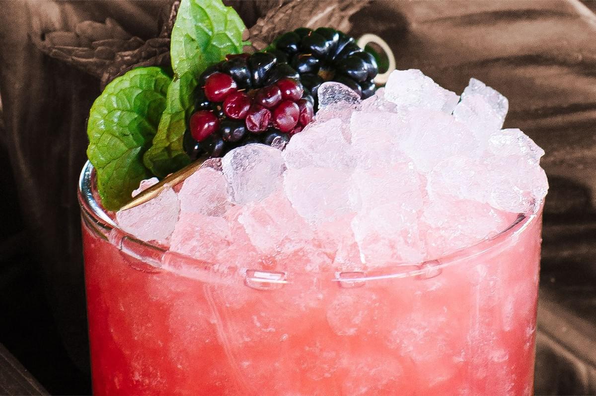 A refreshing pink cocktail in a glass filled with crushed ice. Garnished with mint leaves and blackberries, set against a warm, cozy background. A refreshing pink cocktail in a glass filled with crushed ice. Garnished with mint leaves and blackberries, set against a warm, cozy background.