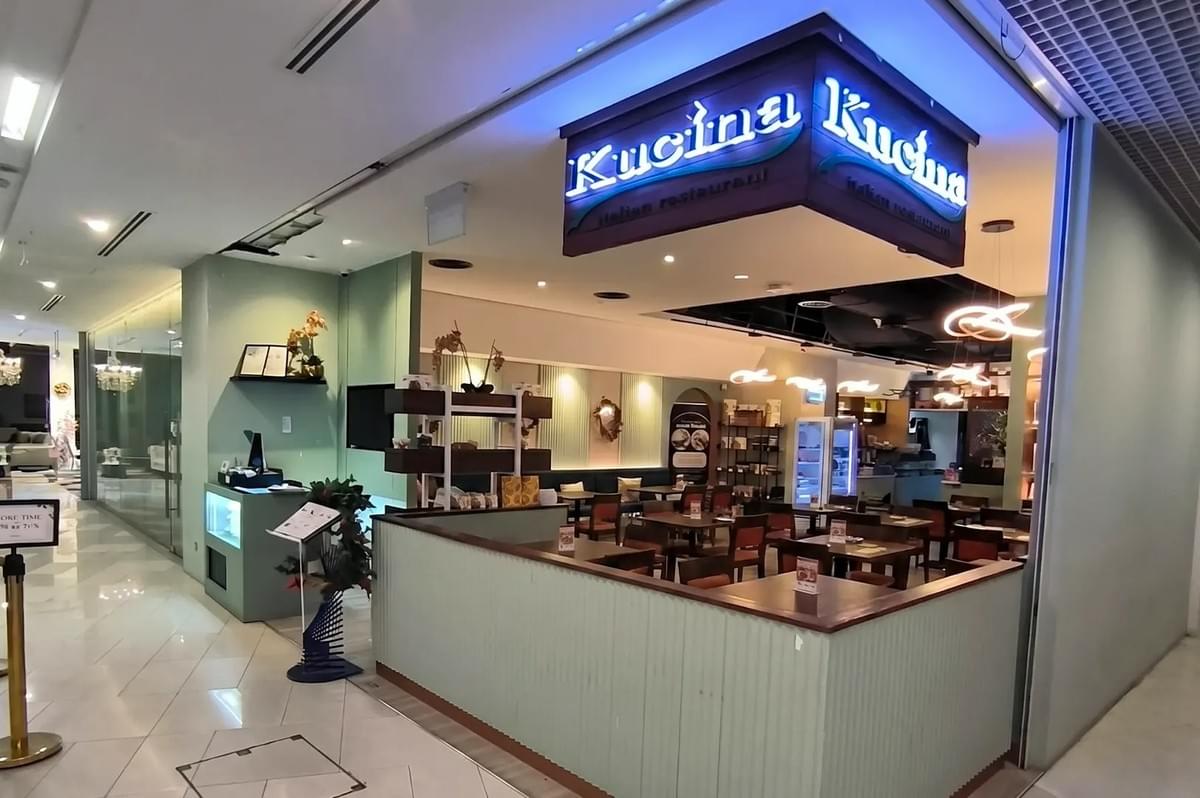 The storefront of Kucina Italian Restaurant located inside a shopping mall, featuring a modern sign, warm lighting, and a dining area with tables and chairs. The storefront of Kucina Italian Restaurant located inside a shopping mall, featuring a modern sign, warm lighting, and a dining area with tables and chairs.
