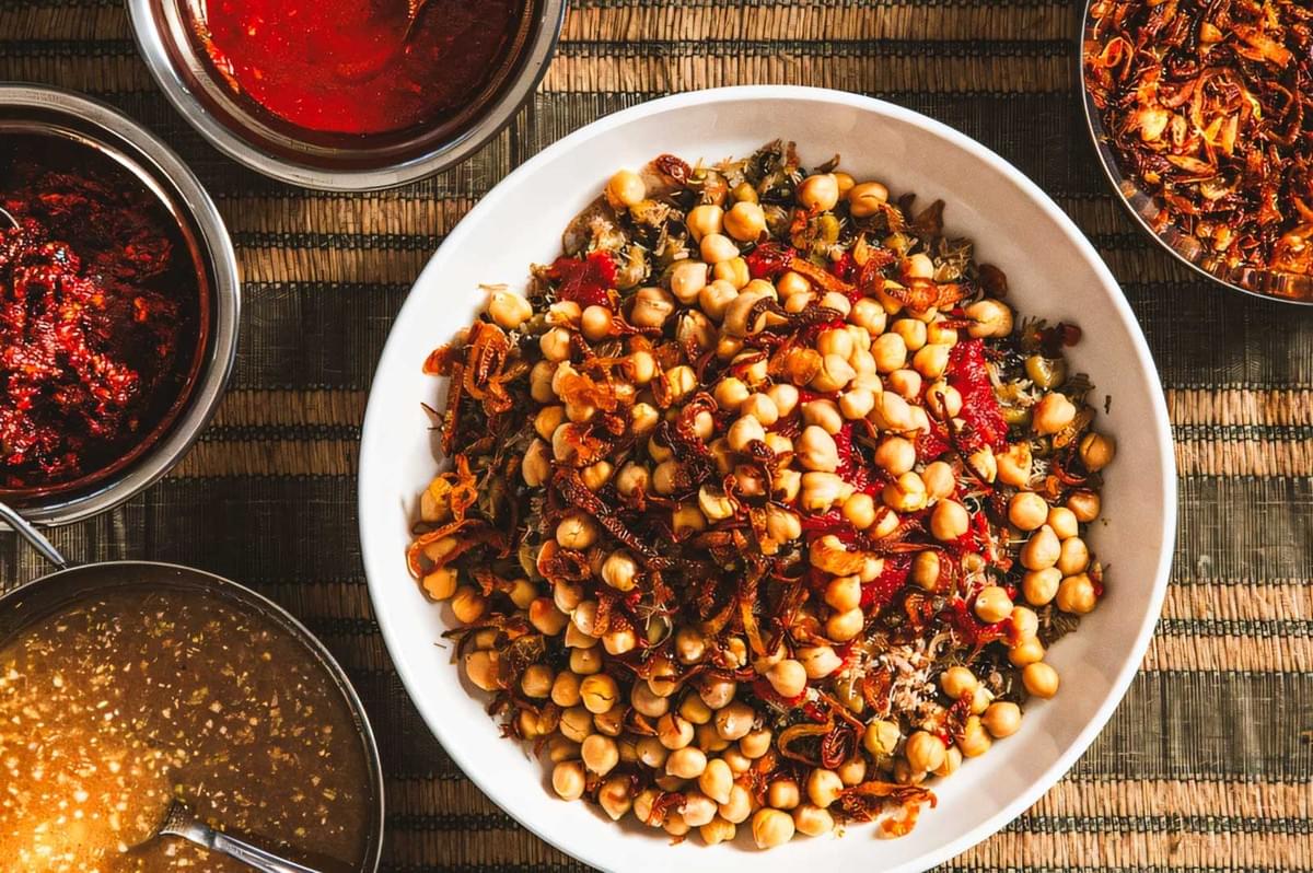 Overhead view of a bowl of Egyptian koshari loaded with chickpeas and fried onions, surrounded by bowls of tomato sauce, vinegar-garlic sauce, and chili paste. Overhead view of a bowl of Egyptian koshari loaded with chickpeas and fried onions, surrounded by bowls of tomato sauce, vinegar-garlic sauce, and chili paste.
