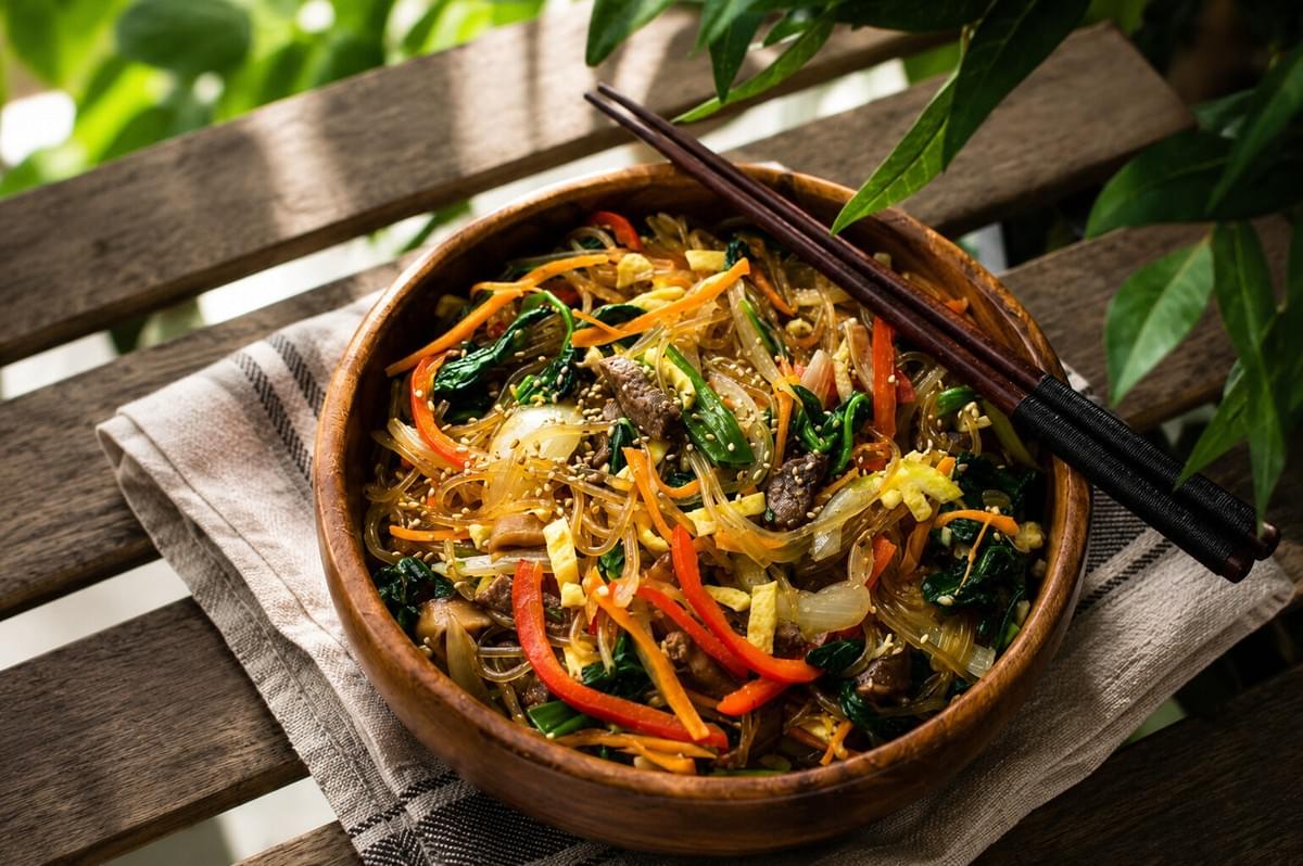 A wooden bowl of colorful Japchae served on a slatted wooden table outdoors. A wooden bowl of colorful Japchae served on a slatted wooden table outdoors.