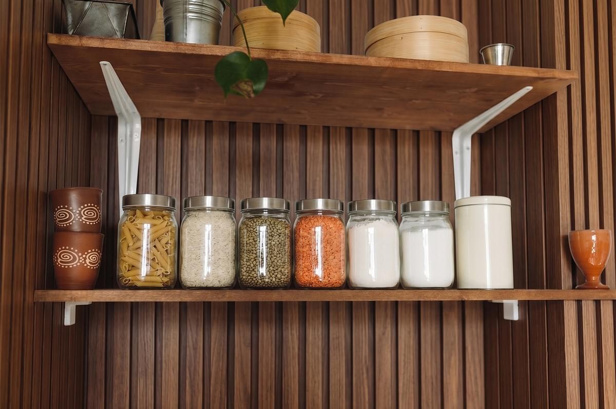 A row of glass pantry jars filled with various grains, legumes, and pasta, arranged neatly on a wooden kitchen shelf with slatted wood paneling in the background. A row of glass pantry jars filled with various grains, legumes, and pasta, arranged neatly on a wooden kitchen shelf with slatted wood paneling in the background.
